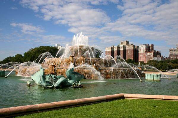 A large, ornate fountain with multiple water jets set in a park area surrounded by green grass; city buildings and a blue sky with clouds are visible in the background. A large, ornate fountain with multiple water jets set in a park area surrounded by green grass; city buildings and a blue sky with clouds are visible in the background.