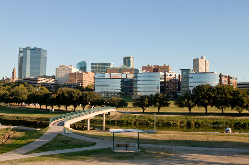 City skyline with modern and high-rise buildings beyond a park with a pathway, a bridge over a small body of water, trees, and an open shelter in the foreground. City skyline with modern and high-rise buildings beyond a park with a pathway, a bridge over a small body of water, trees, and an open shelter in the foreground.